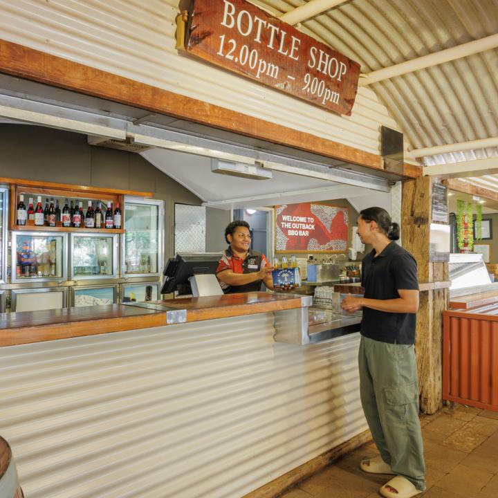 A women is passing a 6 pack of Byron Bay Brewery beers to a customer