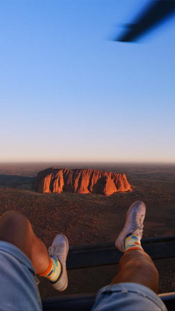 Point Of View of a person sitting on a helicopter dangling their legs over the edge with Uluru down below