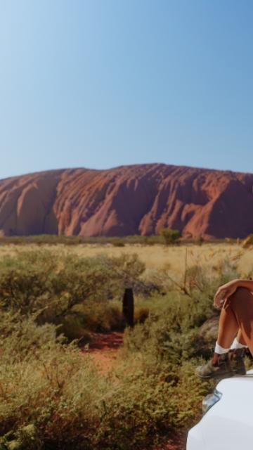 A girl sitting on top of a car with Uluru in the distance